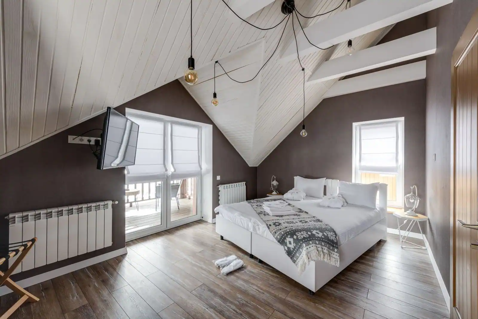Chambre sous combles aux plafonds en pente avec poutres peintes en blanc, un grand lit clair et un parquet en bois