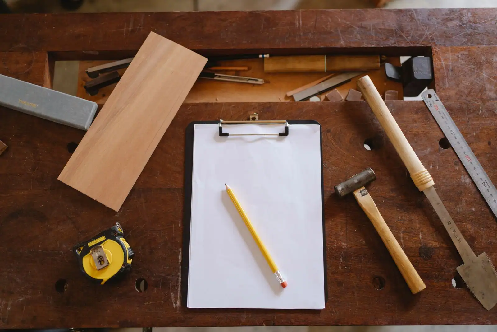 Établi en bois vu de dessus avec un porte-bloc, un crayon et des outils à main (mètre, maillet, scie, réglet)