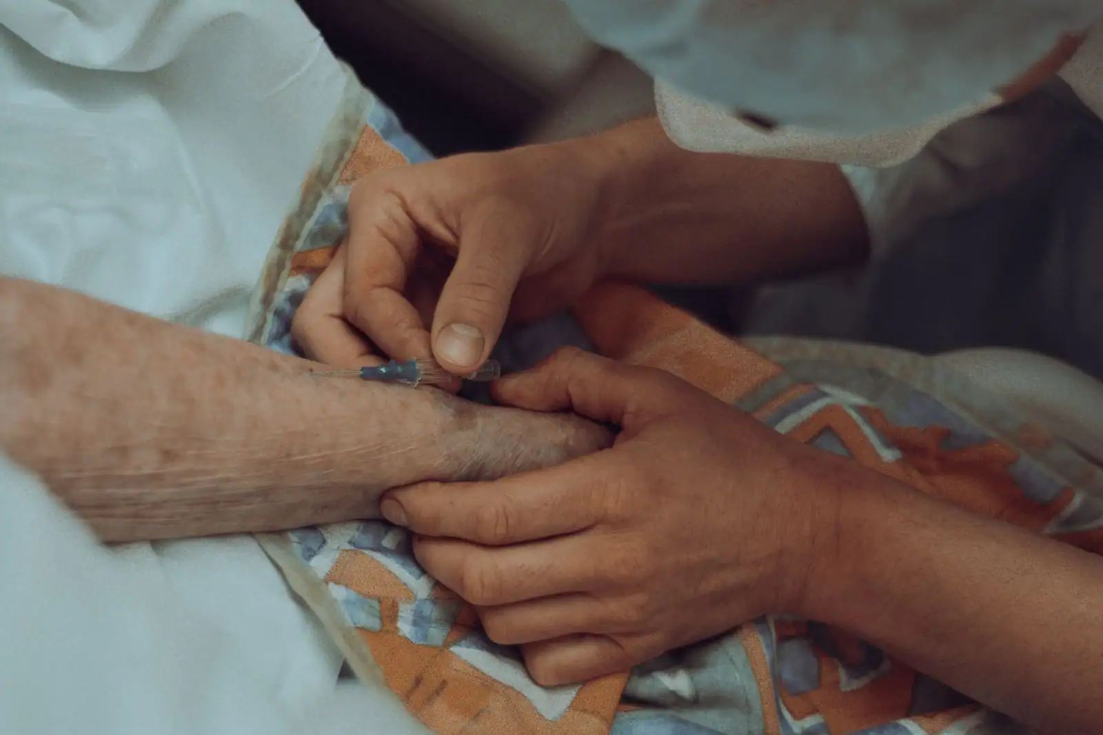 Mains d'une soignante insérant une aiguille dans l'avant-bras d'un patient âgé lors d'un soin.