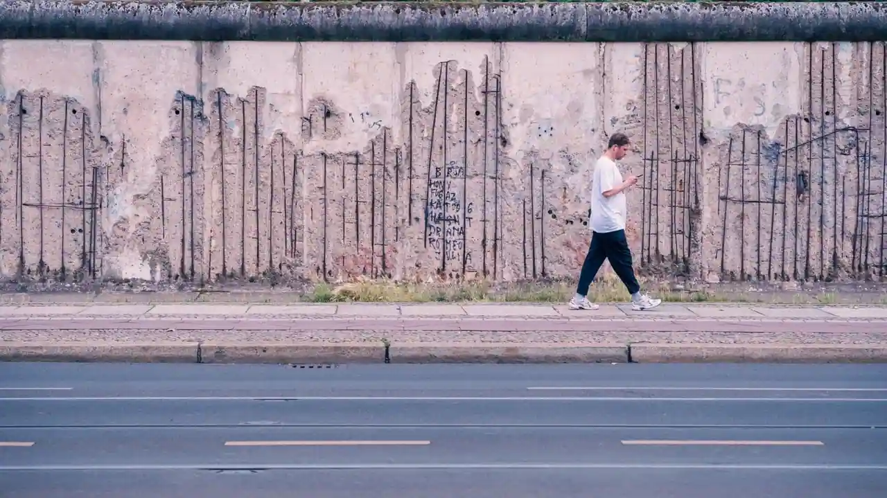 Vestige du mur de Berlin, au béton érodé laissant apparaître les ferraillages, longé par un passant.