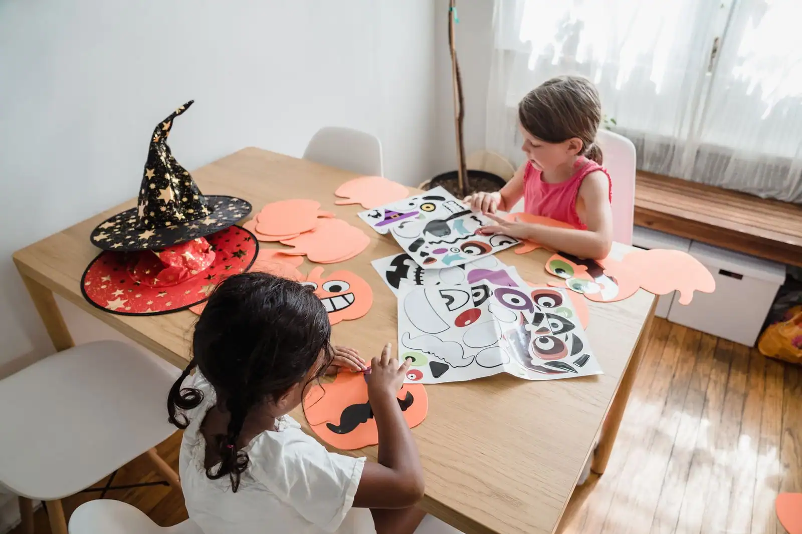 Deux enfants fabriquent des décorations d'Halloween en papier — citrouilles et visages à coller — sur une table en bois.