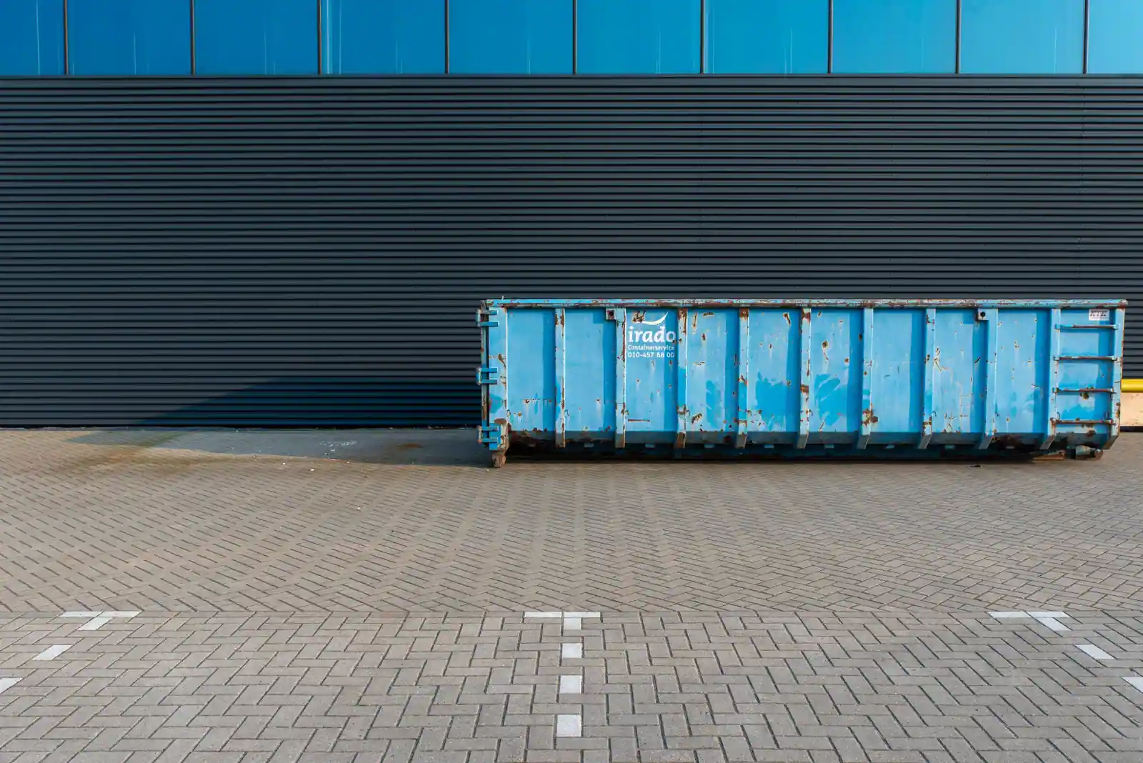 Grande benne de chantier bleue posée sur une aire pavée devant un bâtiment, prête à recevoir les déchets.