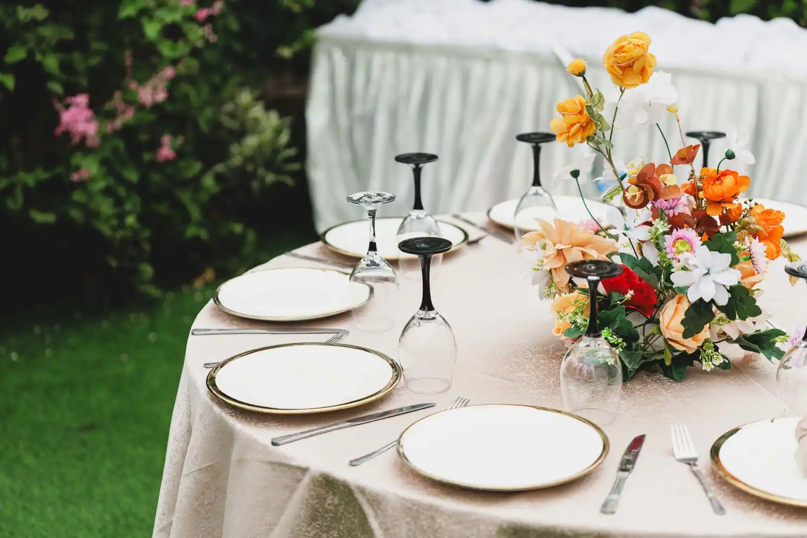 Table de réception dressée en extérieur : assiettes à filet doré, verres à pied et centre de table fleuri.