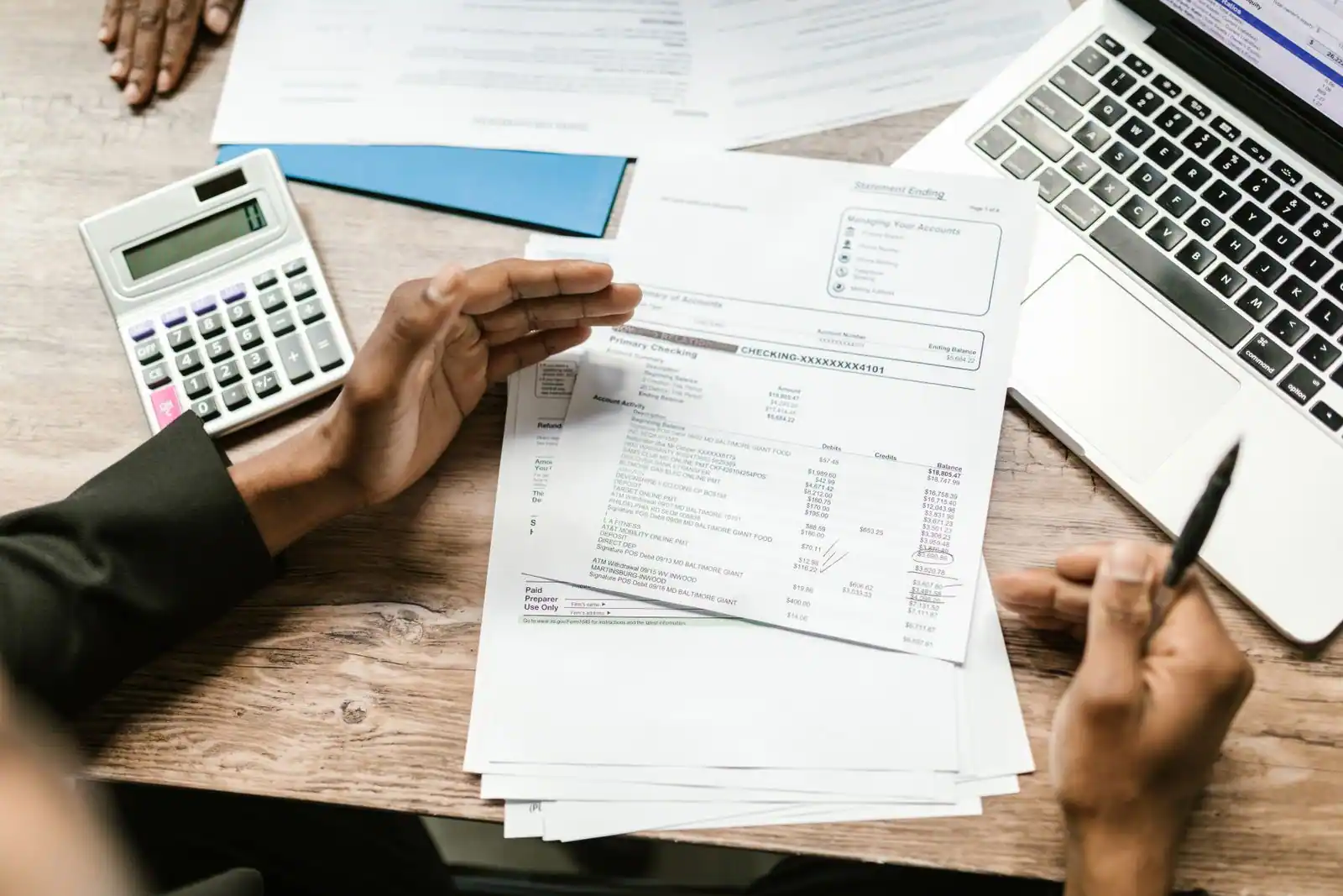 Personne examinant des relevés et documents financiers à un bureau, avec un ordinateur portable et une calculatrice.