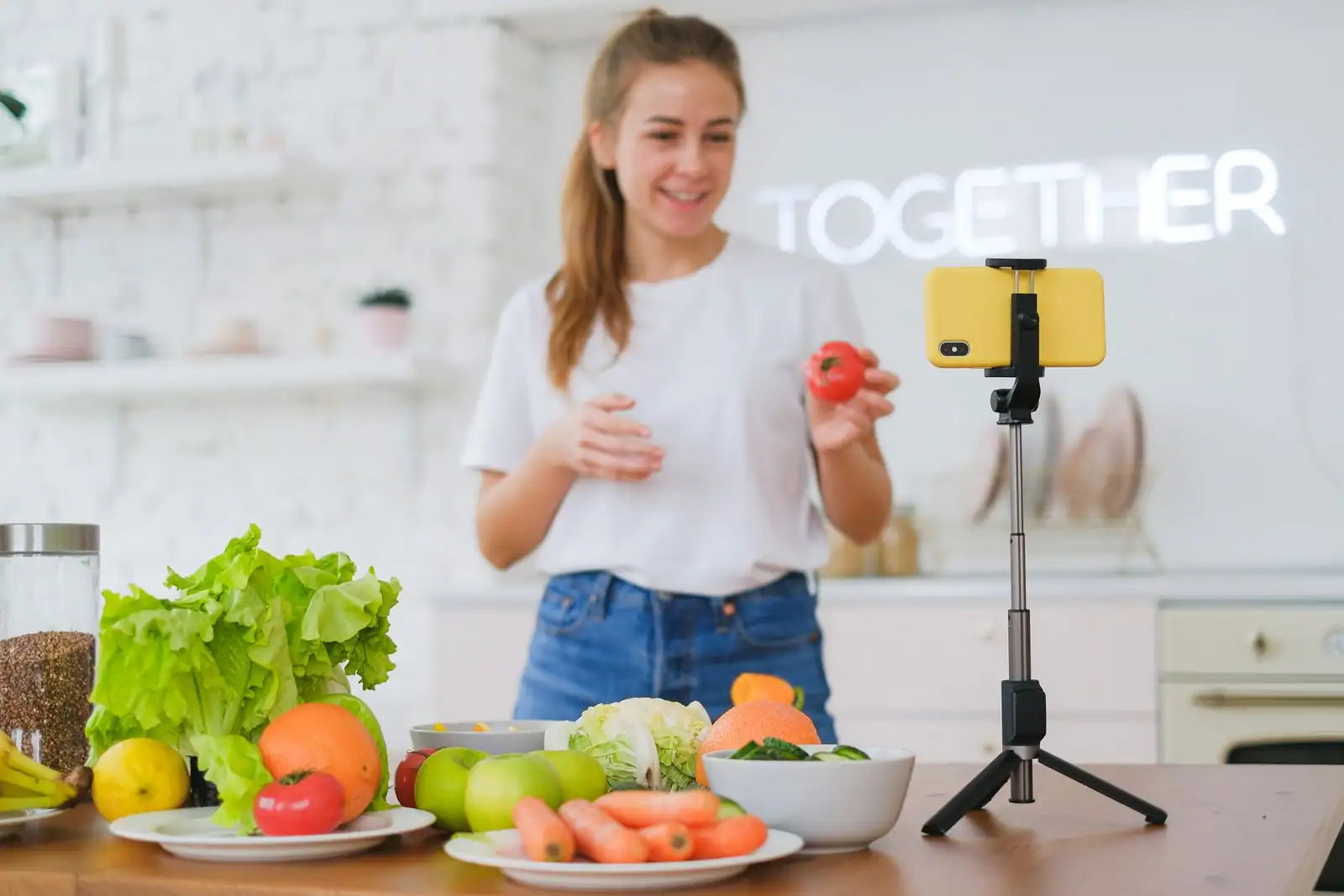 Jeune femme filmant une vidéo avec un smartphone sur trépied dans une cuisine, entourée de légumes