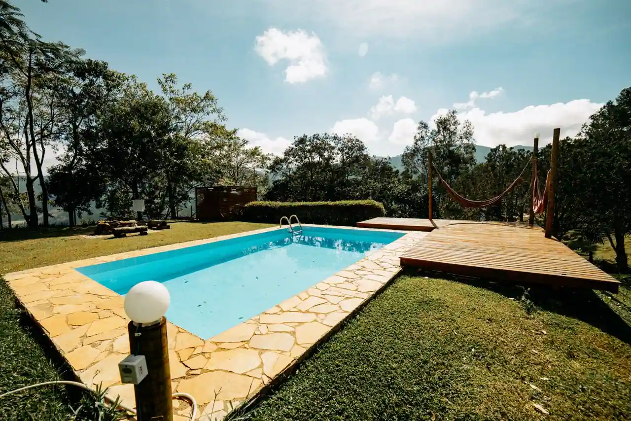 Piscine extérieure à l'eau bleue dans un jardin arboré, bordée de pierre et d'une terrasse en bois, au soleil.