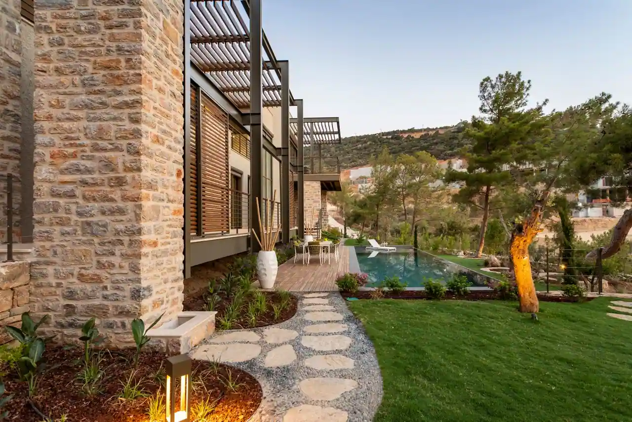Villa en pierre avec pergola, jardin paysager arboré et piscine longeant une allée en pierre naturelle.