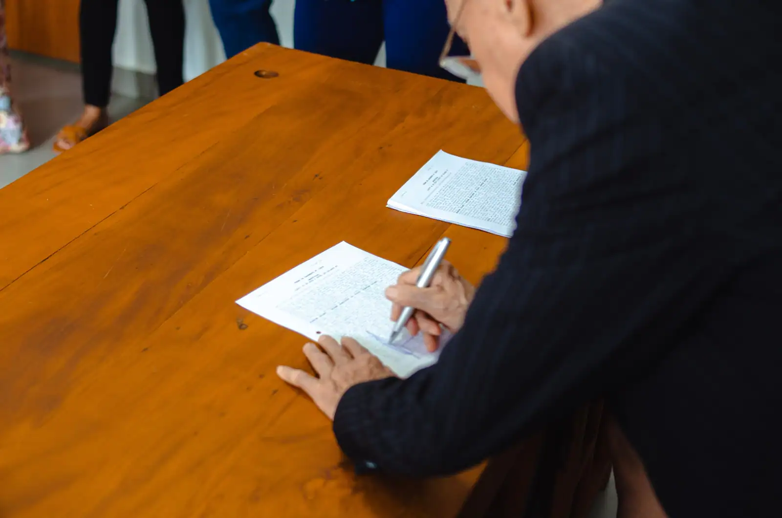 Personne signant un document officiel au stylo sur une grande table en bois, dans un cadre formel