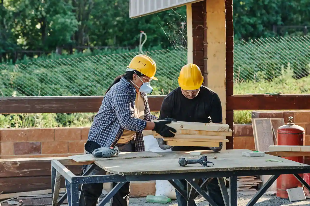 Deux ouvriers casqués et gantés manipulent des planches de bois sur un établi de chantier, outils posés