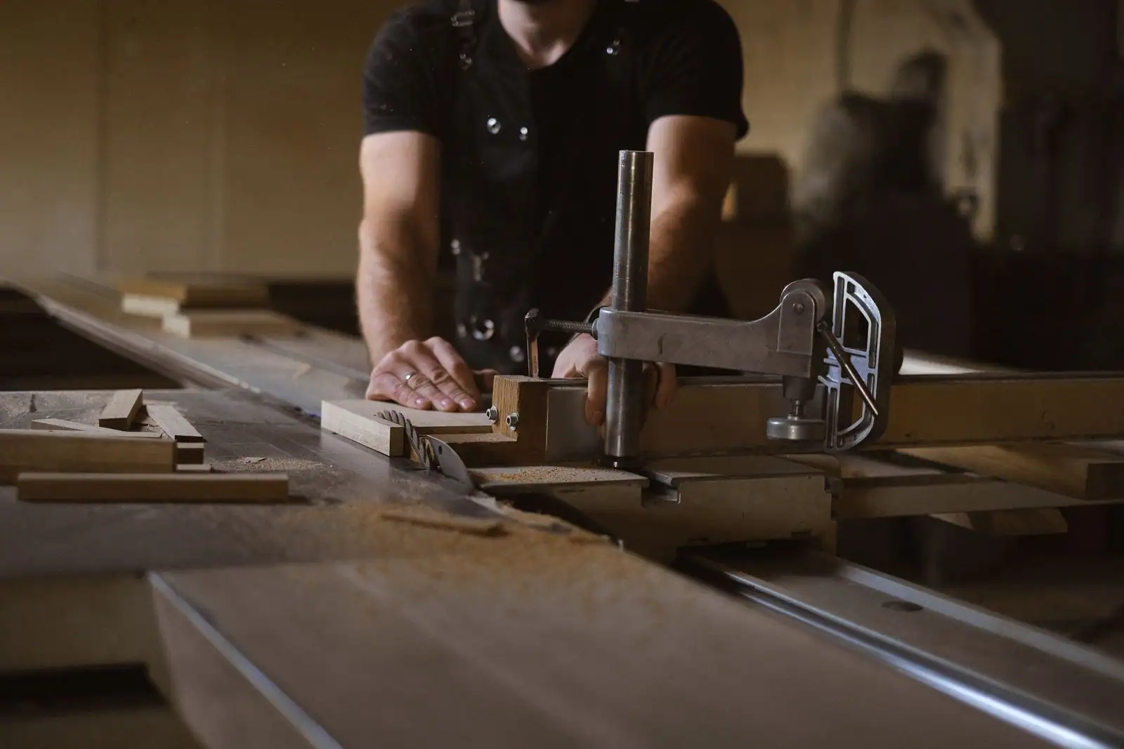 Un menuisier guide une pièce de bois sur la scie de son atelier, maintenue par un serre-joint métallique.